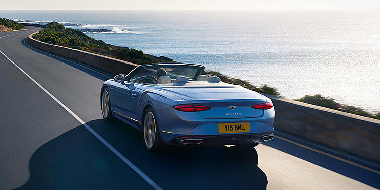 Bentley Bucuresti New Bentley Continental GTC Azure convertible rear view in Light Windsor blue paint with 22 inch ten spoke wheels driving dynamically on an ocean road in the sunshine