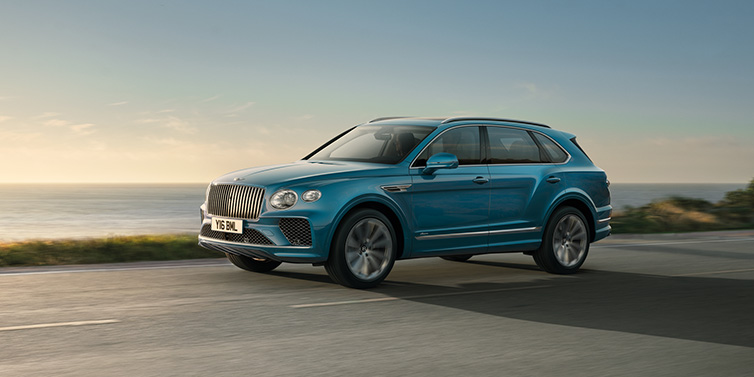 Bentley Bucuresti Bentley Bentayga Azure SUV in Topaz blue paint driving dynamically by the ocean with 22 inch 10 spoke directional wheels