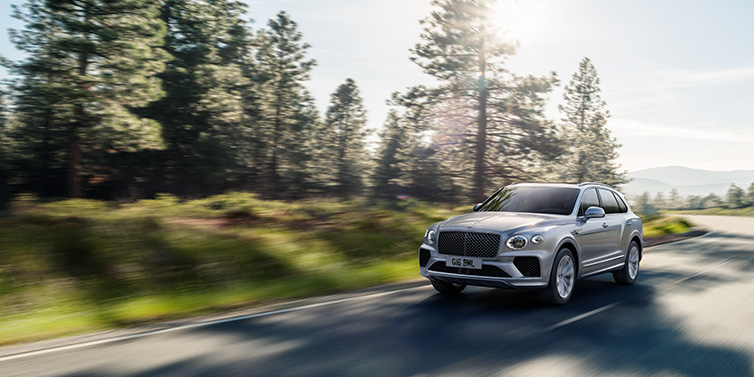 Bentley Bucuresti Bentley Bentayga SUV front three quarter in Moonbeam silver paint driving dynamically along a tree-lined road